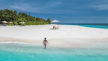 A beach with clear turquoise water, a woman walking in the water, and beach chairs and umbrellas in the background.
