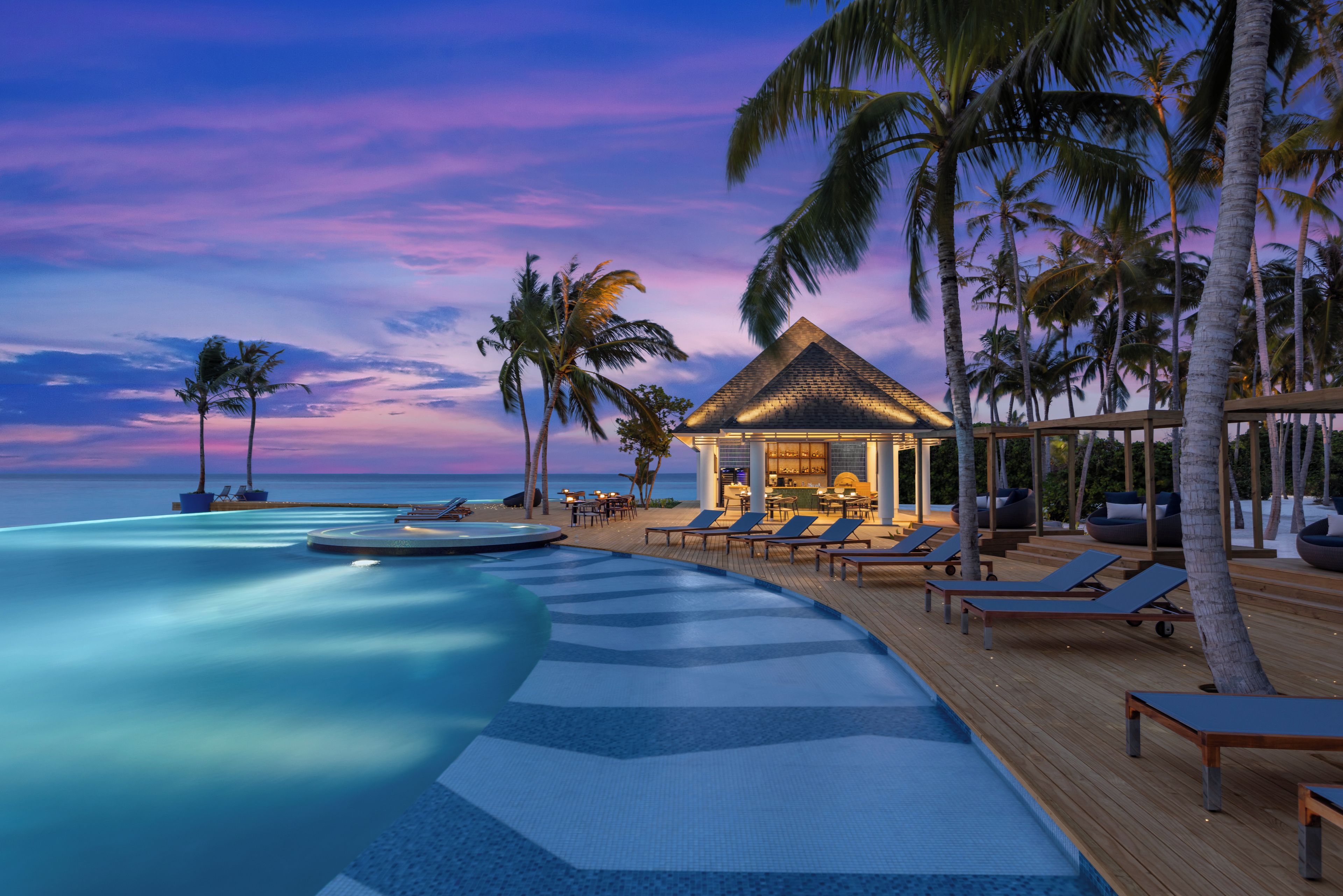 A resort pool area with lounge chairs and a small hut, surrounded by palm trees and overlooking the sea.