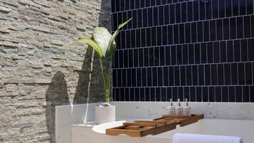 Un baño moderno con pared de piedra, pared con azulejos negros, bañera, estantes de madera y una planta en maceta.
