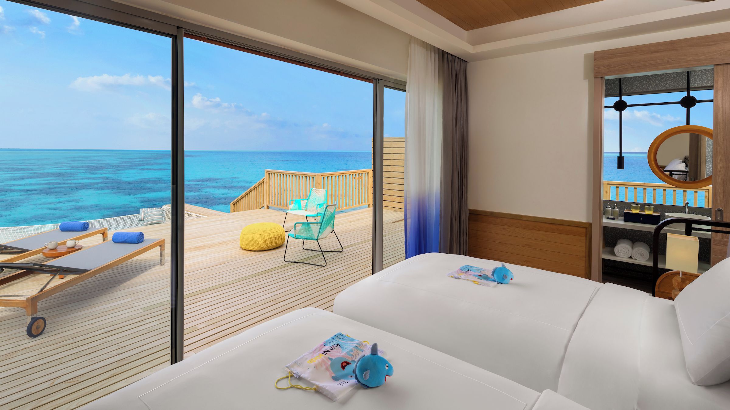 A room with a large window overlooking the ocean, a wooden deck with lounge chairs, and a bathroom with a round mirror and sink.