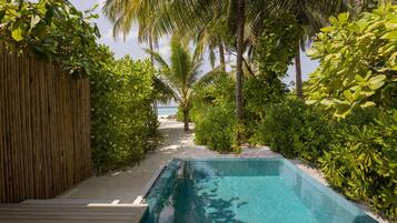 A pool area with a wooden deck, surrounded by lush greenery and palm trees, leading to a beach.