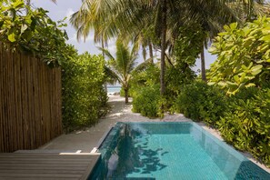 A pool area with a wooden deck, surrounded by lush greenery and palm trees, leading to a beach.