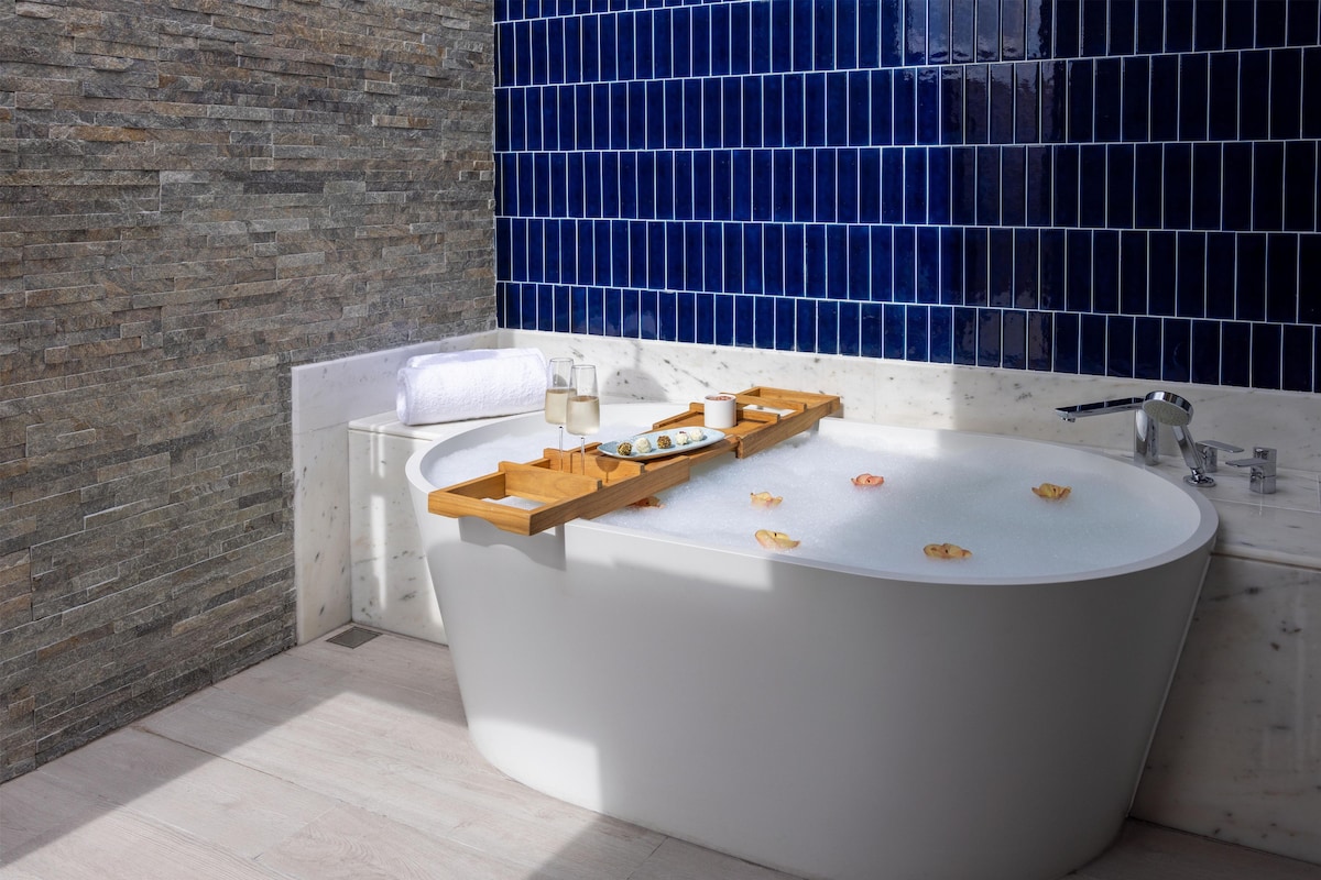 A modern bathroom with a freestanding bathtub, a wooden tray with toiletries, and a marble wall.