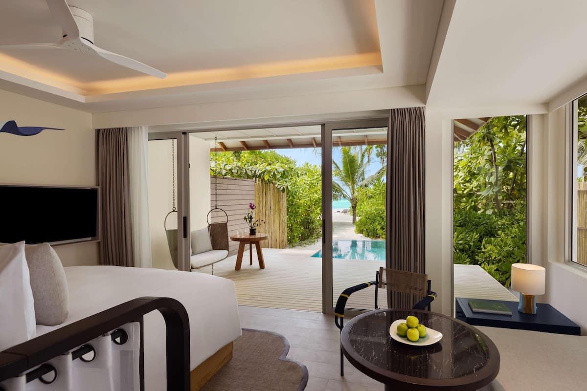 A hotel room with a bed, a TV, a dining table with fruit, a chair, a lamp, and a view of an outdoor pool and greenery.