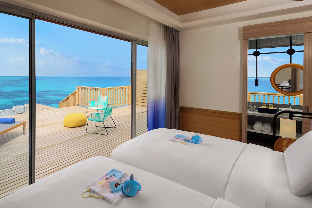 A room with a large window overlooking the ocean, a wooden deck with lounge chairs, and a bathroom with a round mirror and sink.