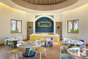 A children's dining area with round tables and colorful chairs, a central play area, and large windows with views of palm trees. - Avani+ Fares Maldives Resort (Fares)