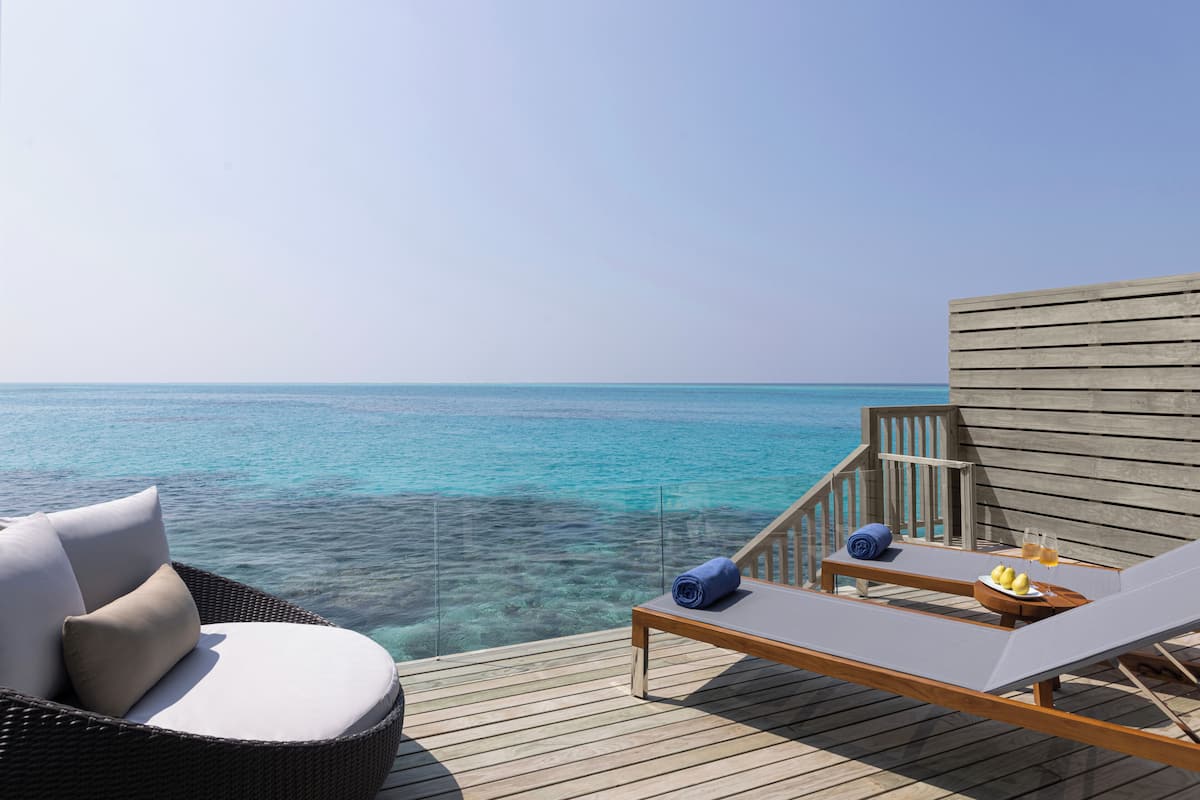 A wooden deck with a lounge chair and a wicker chair, overlooking a clear blue sea.