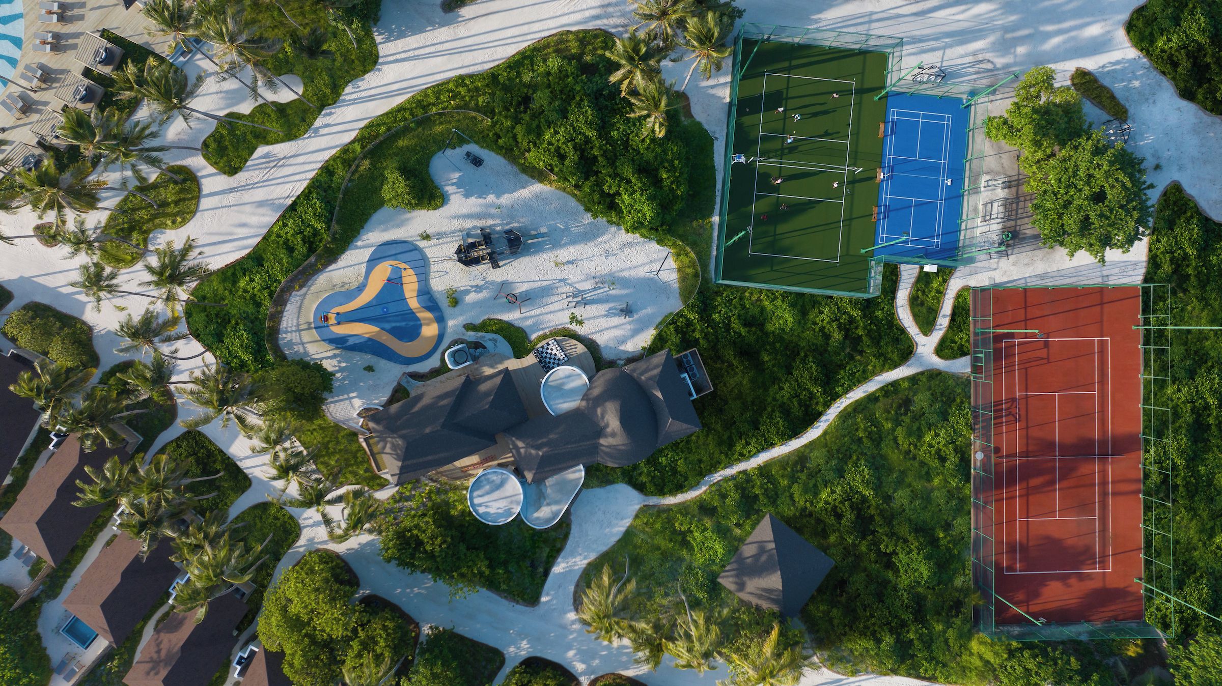 An aerial view of a resort with a sandy beach, a tennis court, and a pool area.