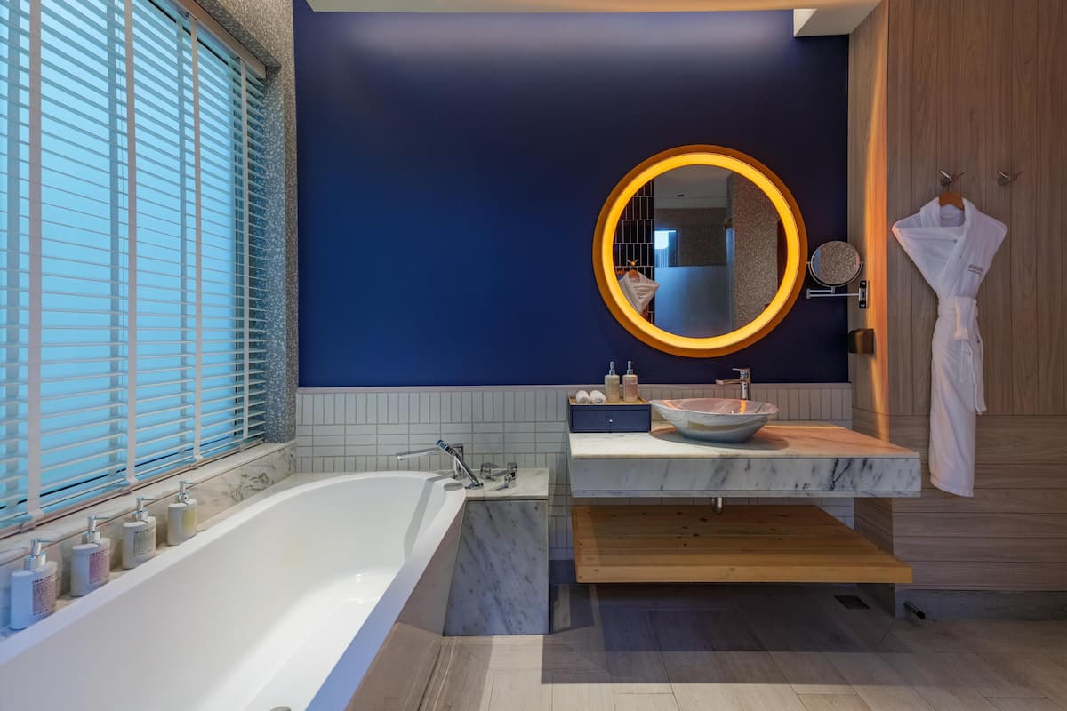 A modern bathroom with a large bathtub, a round mirror with a light, and a wooden vanity.