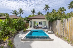 Une piscine entourée d’une terrasse carrelée, un espace salon couvert et une vue sur une maison avec un toit rouge.