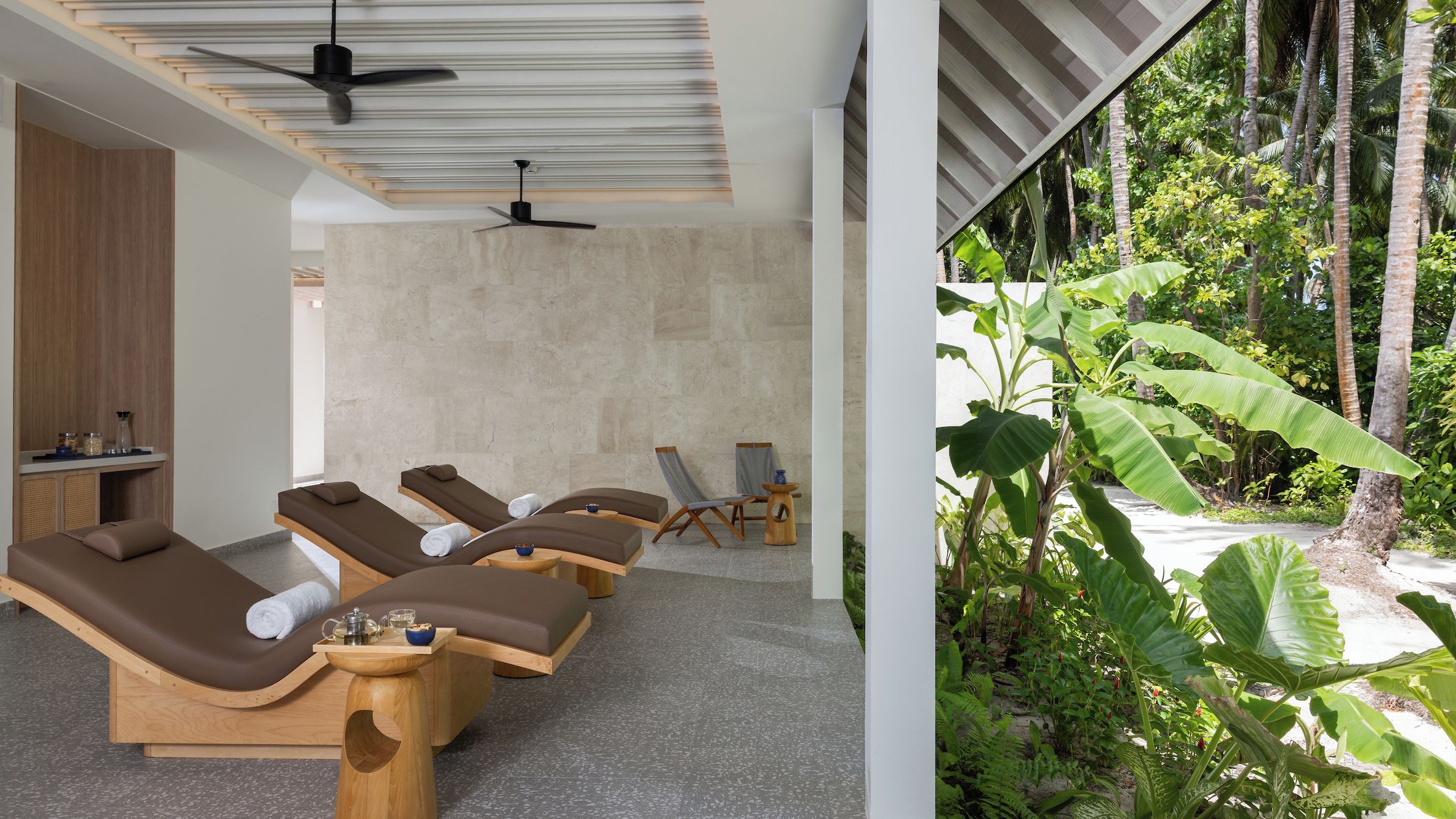 A spa room with multiple sun loungers, a ceiling fan, and a view of a lush tropical garden.