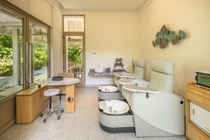 A modern spa room with a large window, a reception desk, multiple seating areas, and a wall-mounted shelf with toiletries. - Avani+ Fares Maldives Resort (Fares)