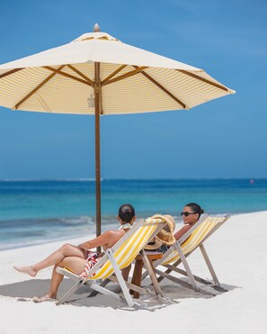 Twee personen zitten op gestreepte strandstoelen onder een gestreepte parasol aan een zandstrand, met een helderblauwe lucht en de oceaan op de achtergrond.