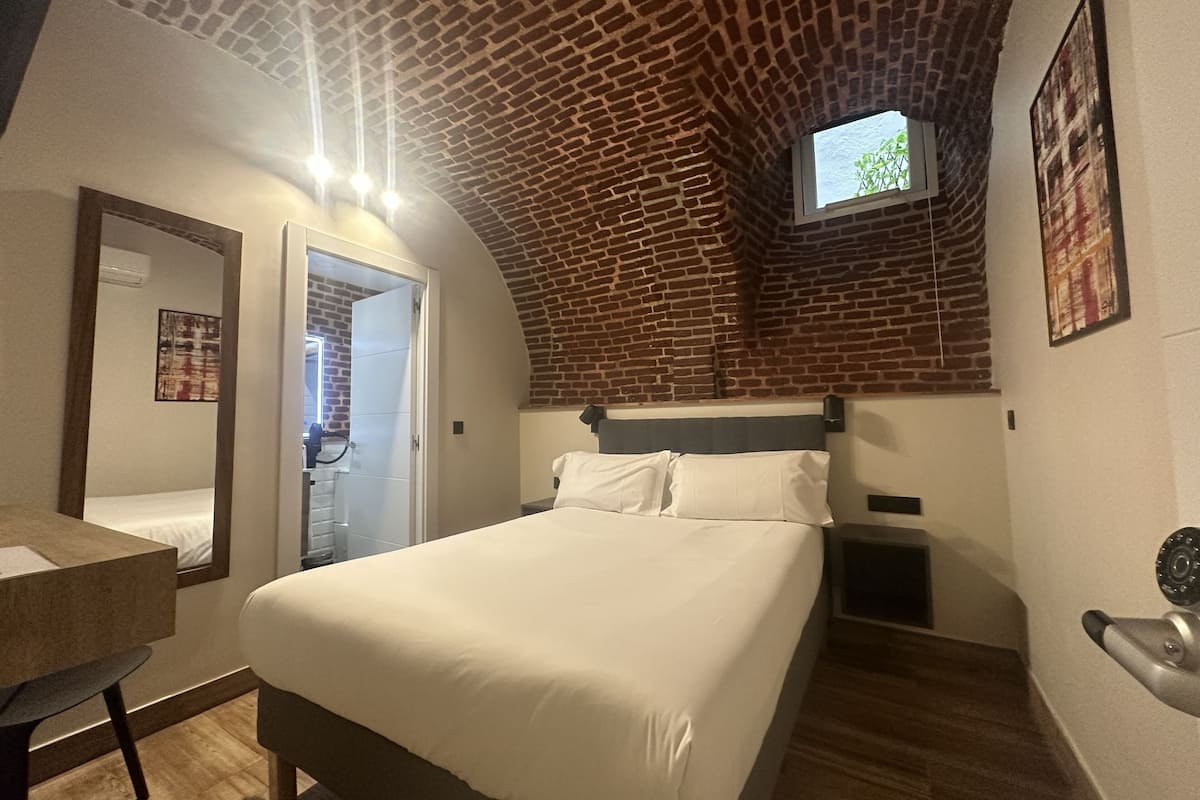 A bedroom with a brick ceiling, a bed, a desk, and a window.