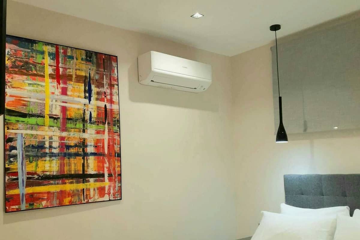A hotel room with a bed, a desk, an air conditioner, a painting, and a pendant light.