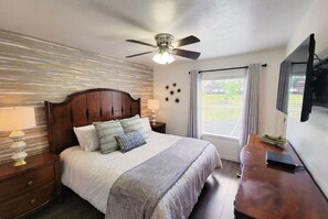 Ein Schlafzimmer mit hölzernem Kopfteil, einem Bett mit weißer Bettwäsche, einem Deckenventilator, einem an der Wand montierten Fernseher und einem Fenster mit Vorhängen.