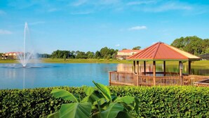 Un gazebo a bordo lago con una fontana, immerso nel verde e con case sullo sfondo.