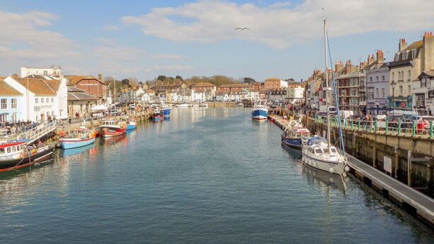 A harbor with boats docked along the waterfront, buildings lining the shore, and a clear sky. - Cove Street Cottage (Weymouth)