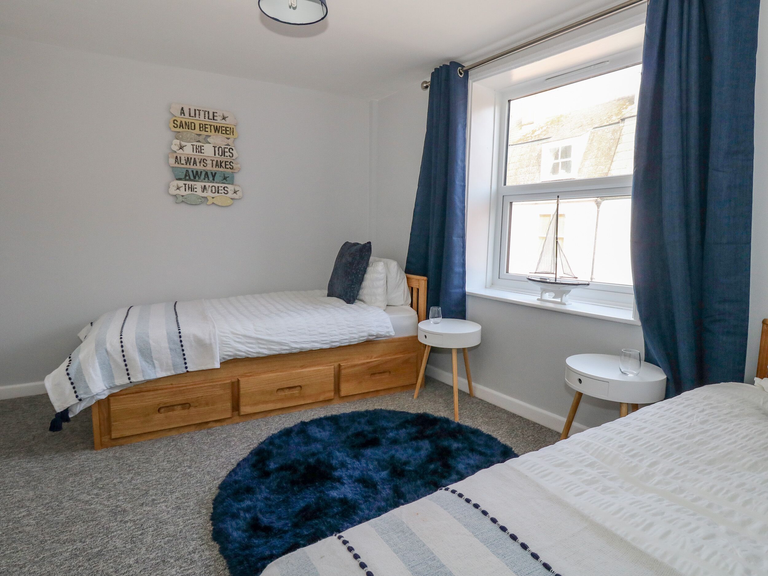 A bedroom with a bed, a small bedside table, a window with curtains, a rug, and a wall decoration.