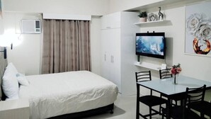 Habitación de hotel con cama, televisor montado en la pared, mesa de comedor con sillas y ventana con cortinas.
