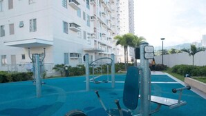 Un área de gimnasio al aire libre con equipo de ejercicio, rodeada por un suelo azul de goma y adyacente a un edificio residencial con balcones.