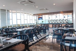 Un restaurante moderno con varias mesas y sillas, una zona de bar central y amplios ventanales que dejan entrar luz natural.