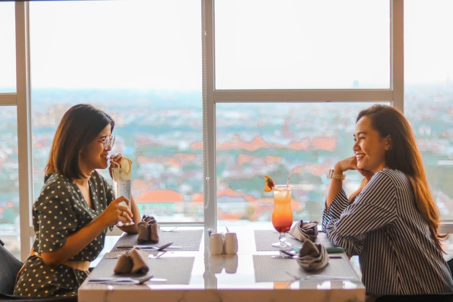 Two individuals seated at a table with a view of a cityscape through large windows.