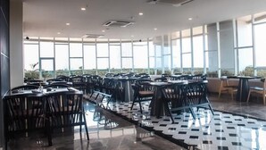 A spacious indoor dining area with multiple tables and chairs, large windows providing natural light, and a checkered floor pattern. - BeSS Mansion Hotel Surabaya (Wonocolo)