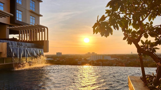 A rooftop pool with a waterfall feature, overlooking a cityscape during sunset. - BeSS Mansion Hotel Surabaya (Wonocolo)
