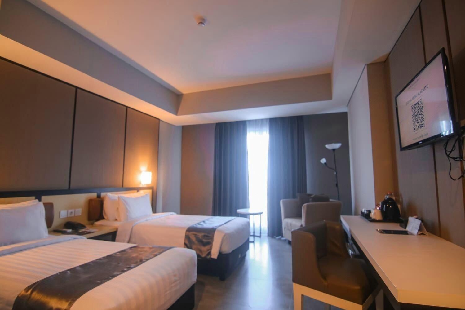 A hotel room with two beds, a desk, a chair, a lamp, and a window with curtains.