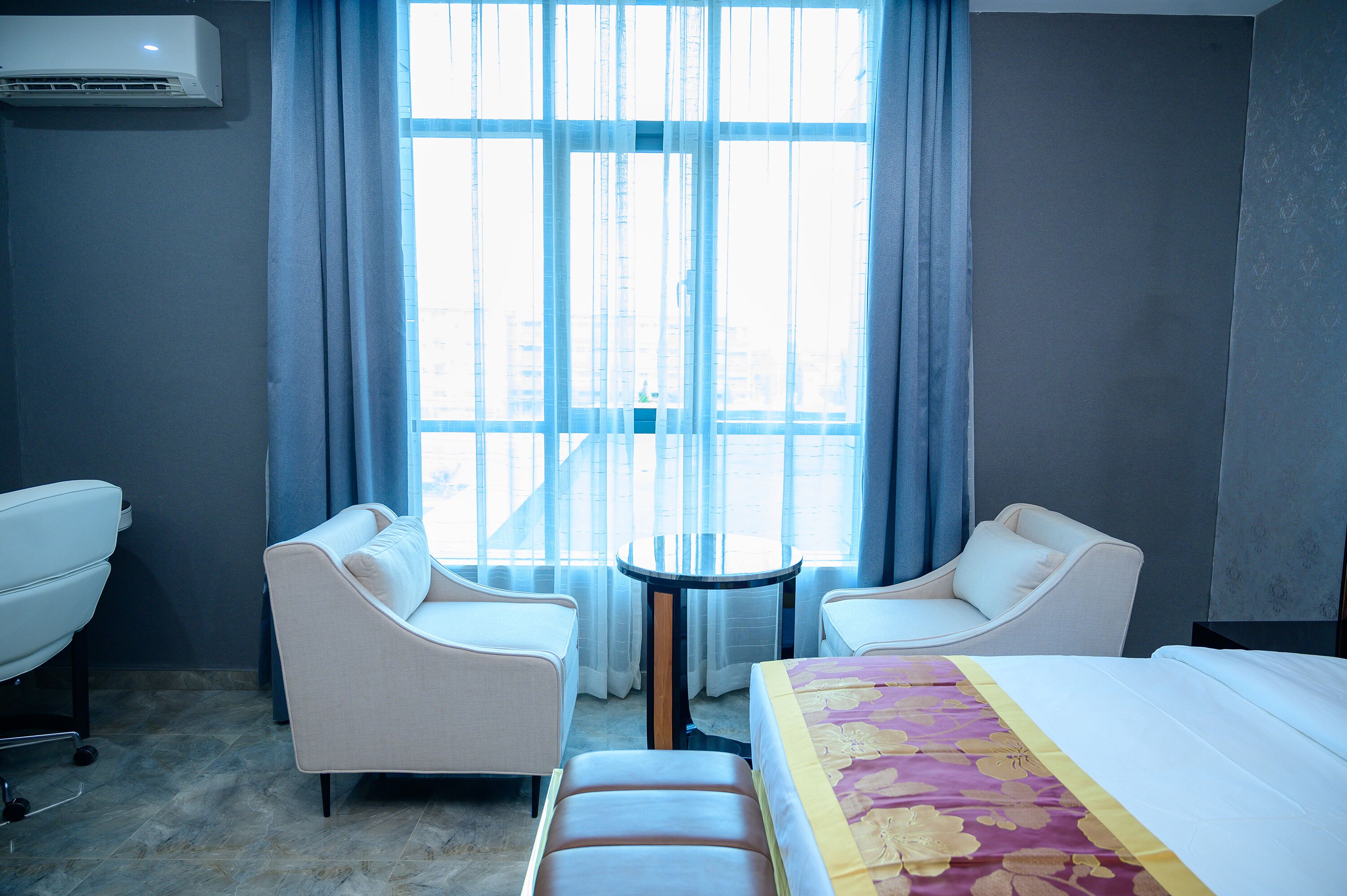A hotel room with a bed, two white armchairs, a small round table, and a window with curtains.