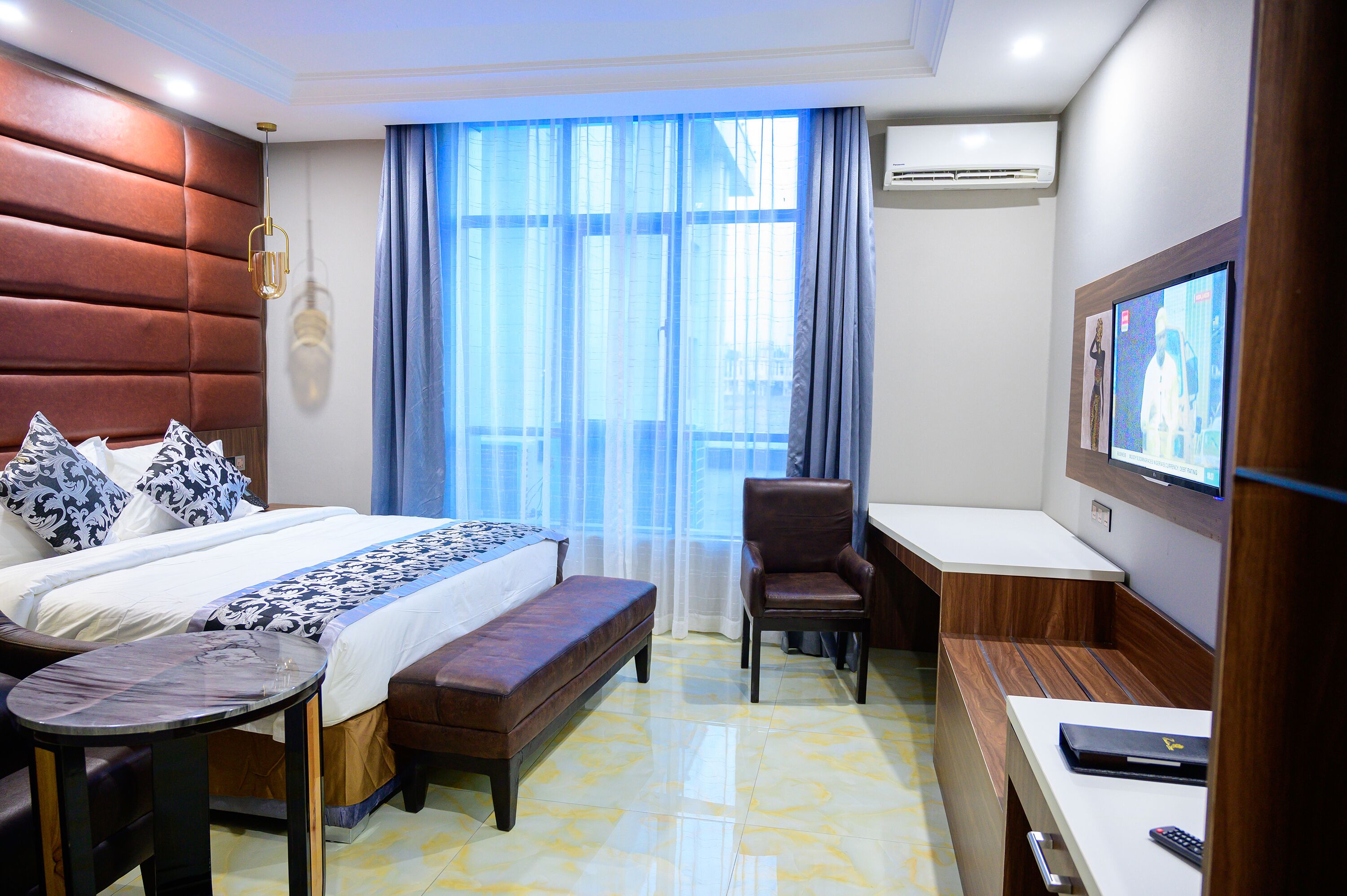 Una habitación de hotel con una cama grande, un banco, un escritorio, una silla, un televisor y un ventanal con cortinas.