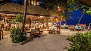 An outdoor restaurant with tables and chairs arranged under a covered area, surrounded by tropical plants and palm trees. - Fair House Beach Resort & Hotel (Koh Samui)