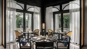 A dining area with round tables and chairs, large windows with curtains, and a view of greenery outside. - TIA Wellness Resort (Da Nang Municipality)