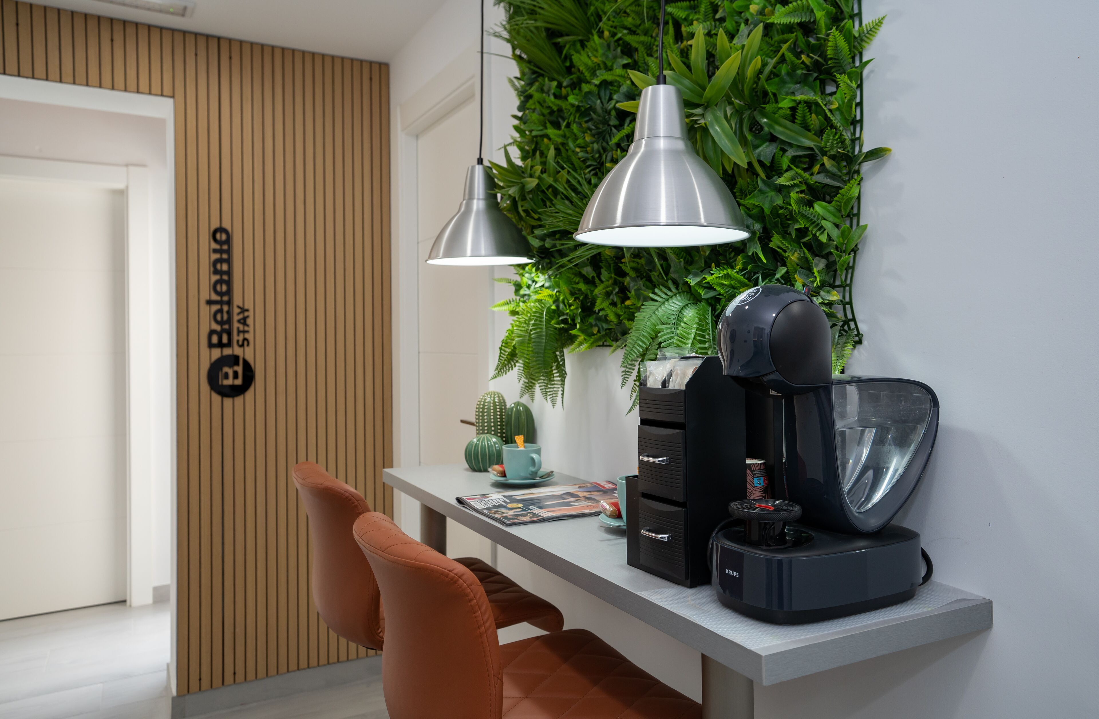Un espacio de oficina moderno con un escritorio, dos lámparas colgantes, una cafetera y una planta en la pared.