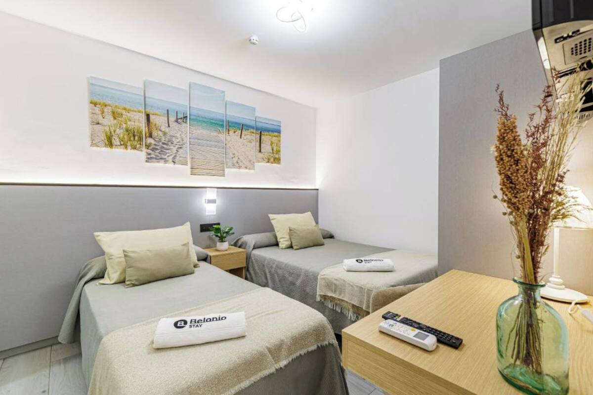 Ett hotellrum med två sängar, ett skrivbord med en fjärrkontroll, en vas med torkade växter och en vägg dekorerad med konstverk med strandtema.