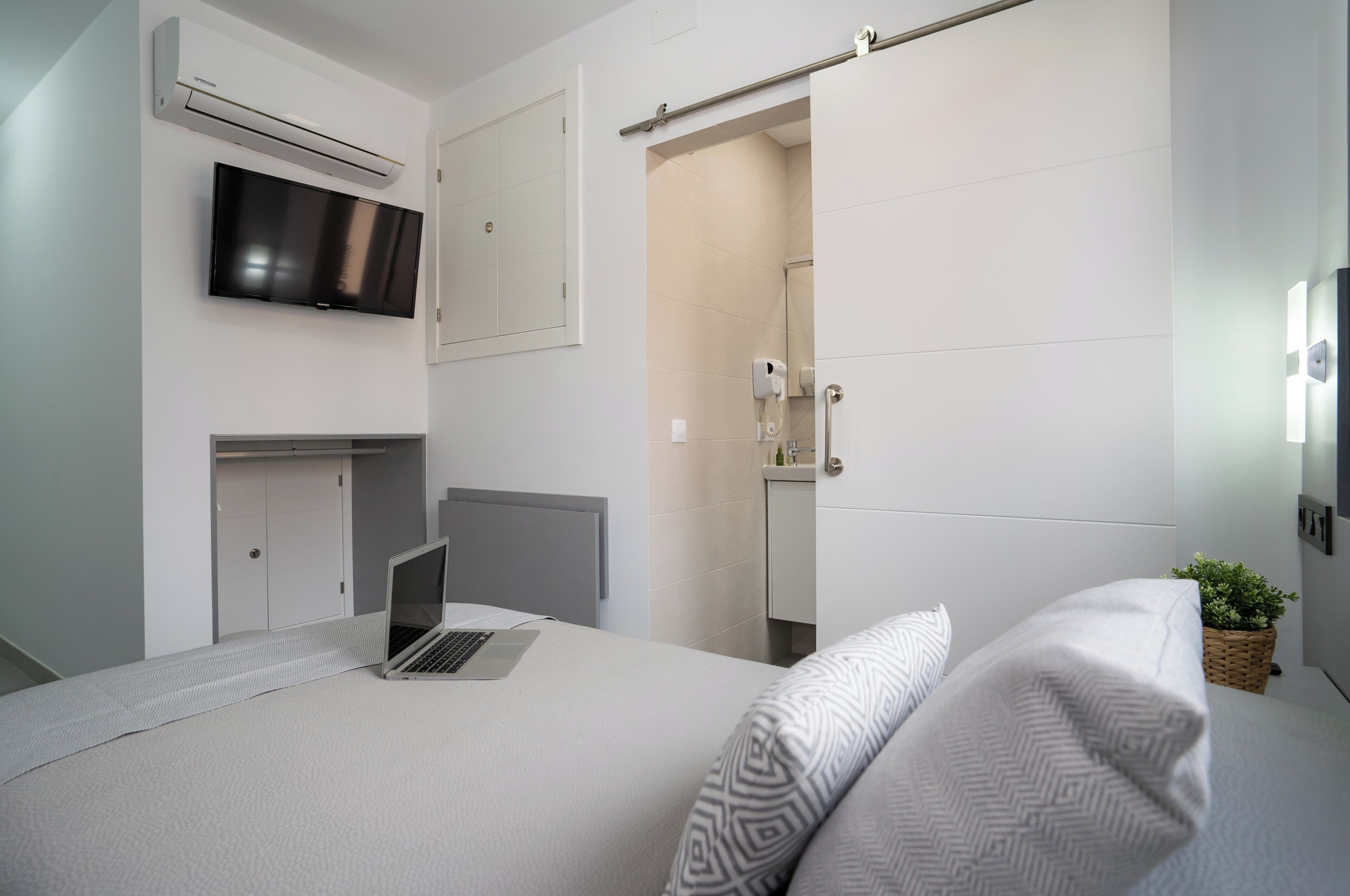 A modern bedroom with a bed, a laptop on the side, a TV mounted above the bed, and a shower area visible through an open door.
