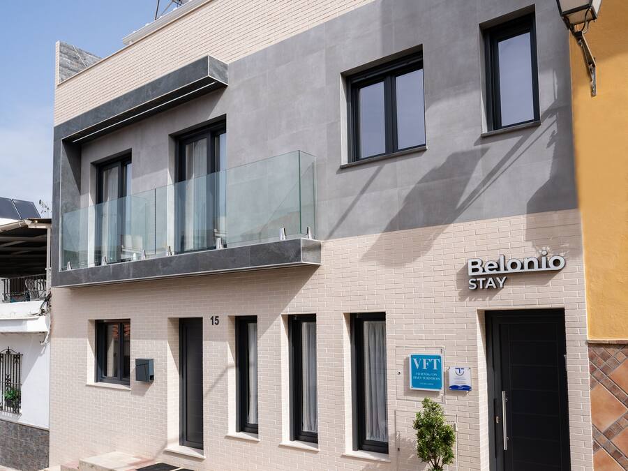 Un edificio de dos pisos con un letrero que dice "Belolio Stay". Cuenta con un balcón con barandillas de vidrio, varias ventanas y una puerta con un letrero que indica servicio de Valet Parking (VFT).