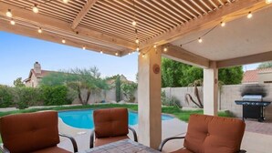 An outdoor patio with a pool, two cushioned chairs, a table with a bowl of snacks, and a grill. - Red Mountain Ranch home, BBQ, pool (Mesa)