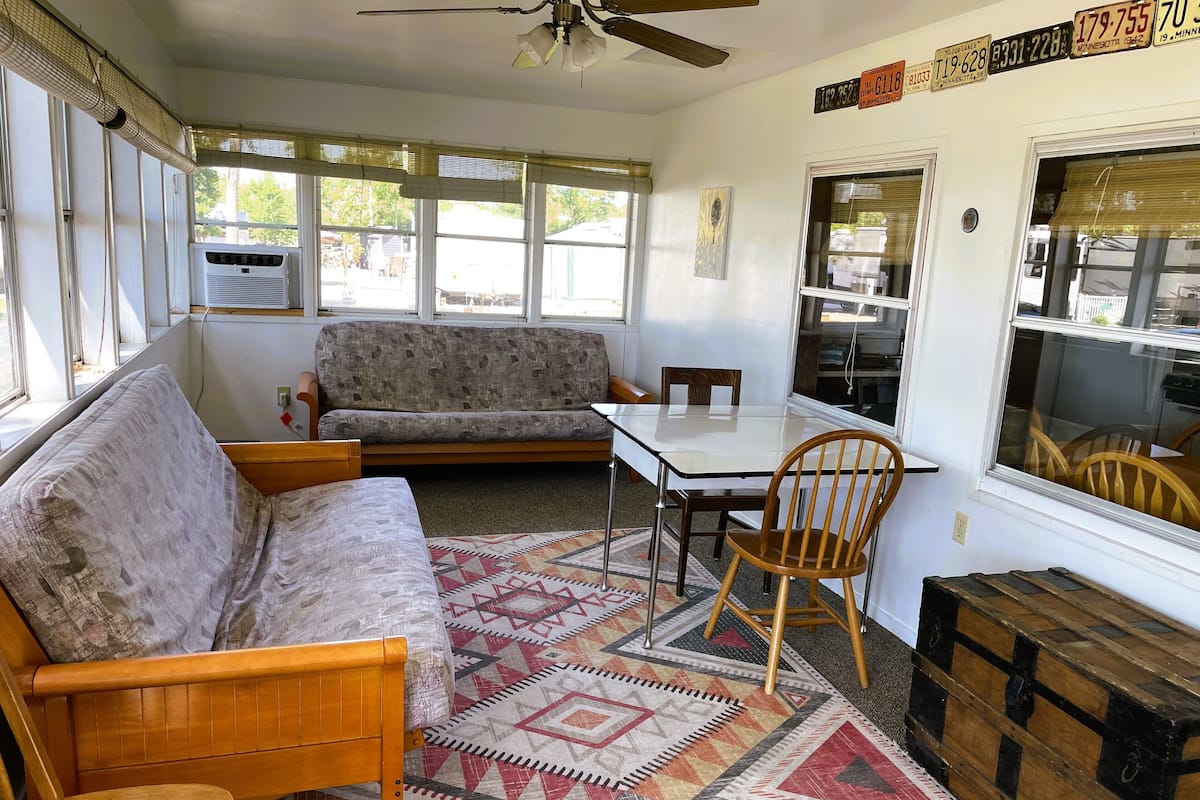 A cozy room with a wooden bed, a sofa, a dining table with chairs, a patterned rug, and a large window with blinds.