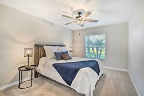 Un dormitorio con cama, ventilador de techo, mesita de noche con lámpara y ventana con persianas.