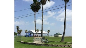 Un panneau indiquant l’entrée du parc Bayshore, situé dans le comté de Galveston, au Texas. On y voit une zone herbeuse, des palmiers et, au fond, un bâtiment doté d’un toit végétalisé.