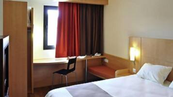 Habitació d'hotel amb llit, escriptori, cadira i finestra amb cortines.