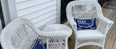 Two wicker chairs with cushions on a wooden deck, adjacent to a white building with horizontal siding.