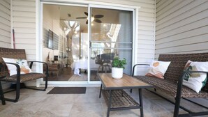 An outdoor patio with two wicker chairs and a wooden table, a sliding glass door leading to a room, and a ceiling fan. - The Nomad at Gables | King Bed • Pool • Garage (Gainesville)