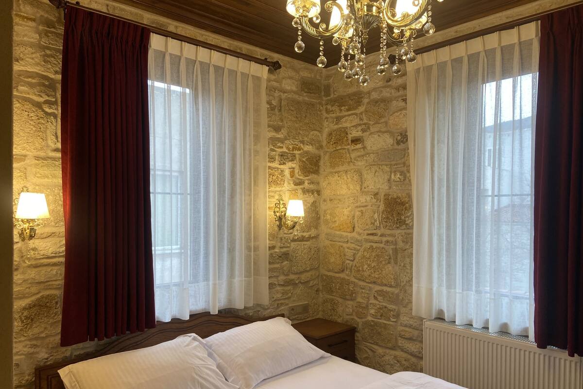 Quarto com parede de pedra, cama com lençóis brancos, duas janelas com cortinas leves e um candelabro.