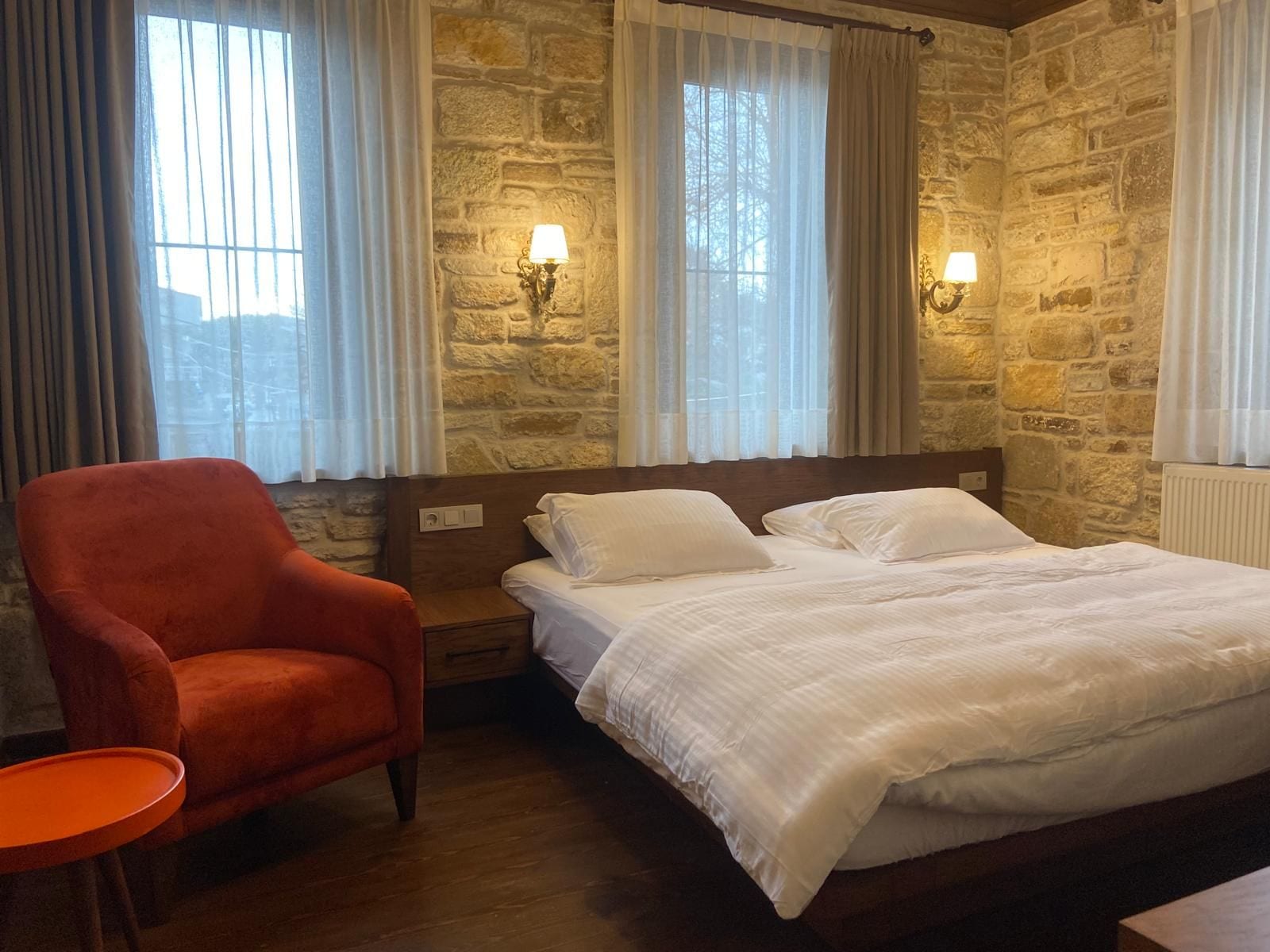 A hotel room with a stone wall, a bed with white bedding, a red armchair, and two wall-mounted lamps.
