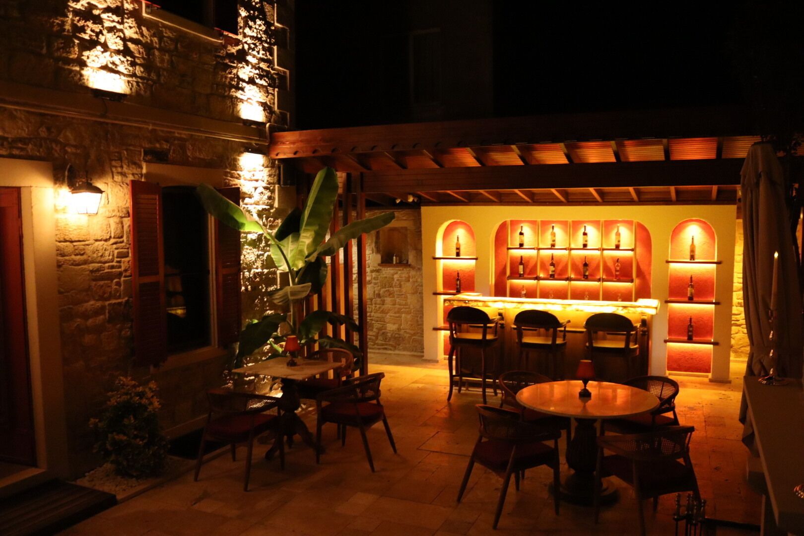 An outdoor patio area with stone walls, wooden furniture, and a bar with illuminated shelves.
