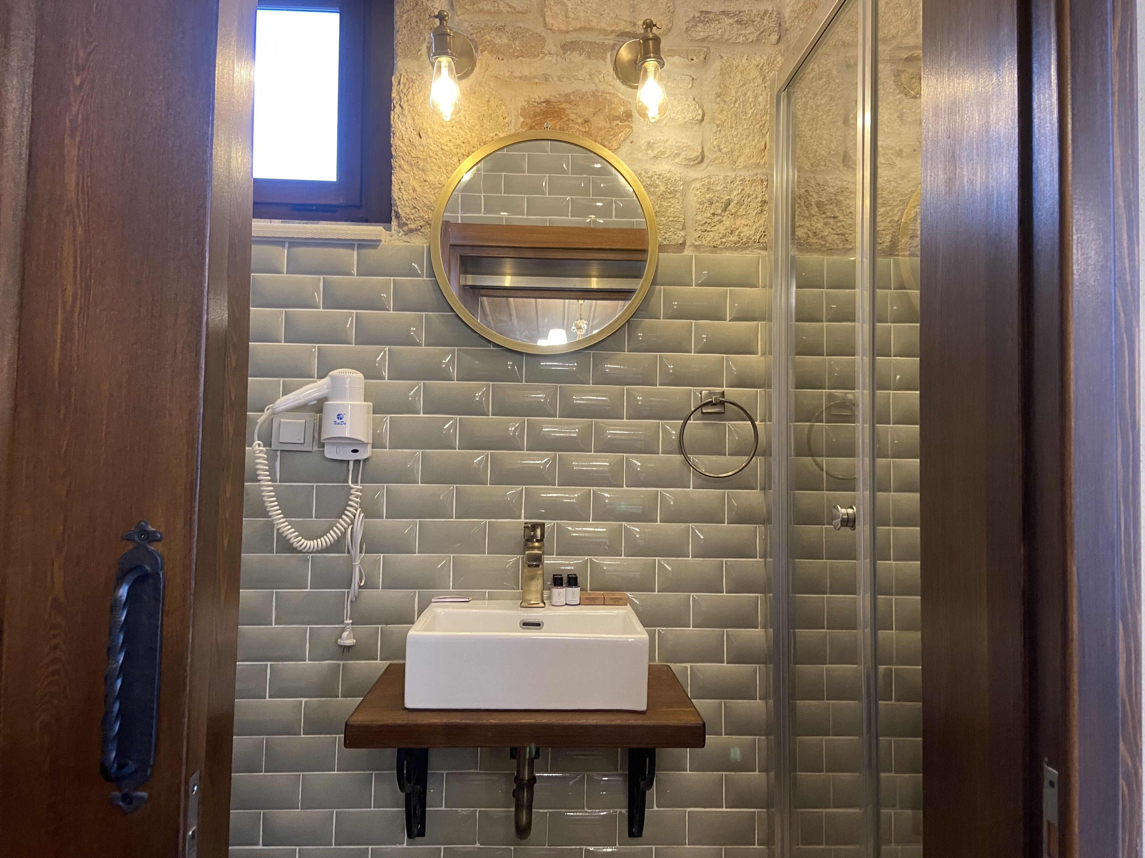 A bathroom with a wooden vanity, a round mirror, a towel ring, and a hairdryer on the wall.