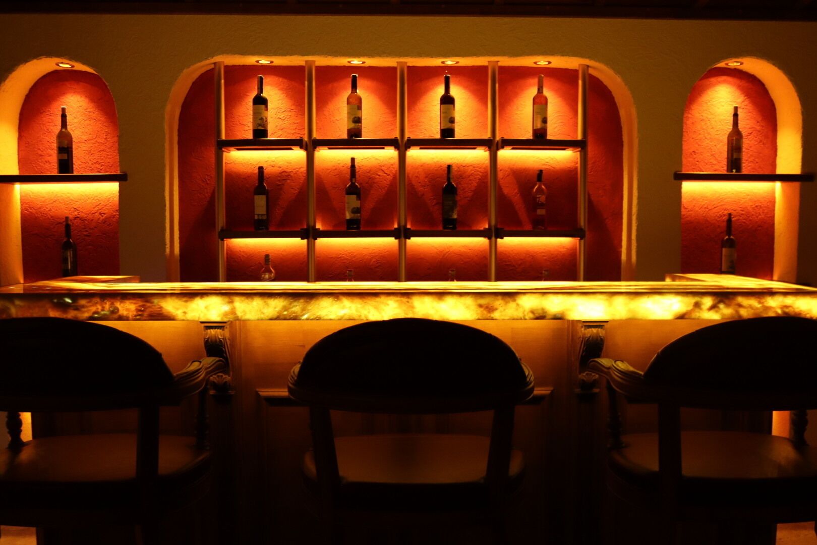 Una zona de bar con una barra alargada y numerosas botellas expuestas en estantes iluminados.